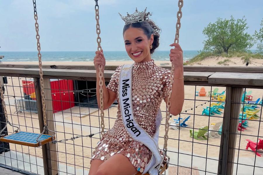 Miss Michigan sitting on a swing on a beachfront deck, wearing a crown and a sequined dress, smiling at the camera.