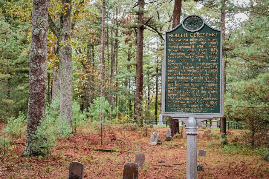 Sign at the entrance of Mouth Cemetery surrounded by trees and graves, indicating historical information about the site.