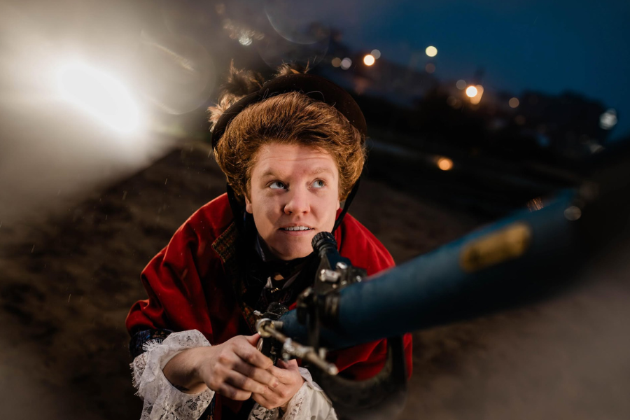 Person in vintage attire and red cloak looks determined while holding a metal pipe. A glowing light and blurred cityscape provide a dramatic backdrop.