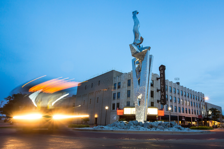 Tall, abstract metal sculpture with reflective surfaces stands before the historic Frauenthal Theater at dusk, with blurred car lights in motion.