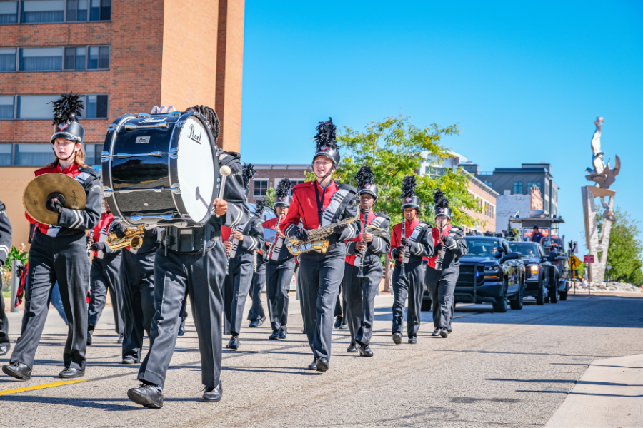 Marching band members in red uniforms performing in a parade on a sunny day, with spectators and buildings in the background.