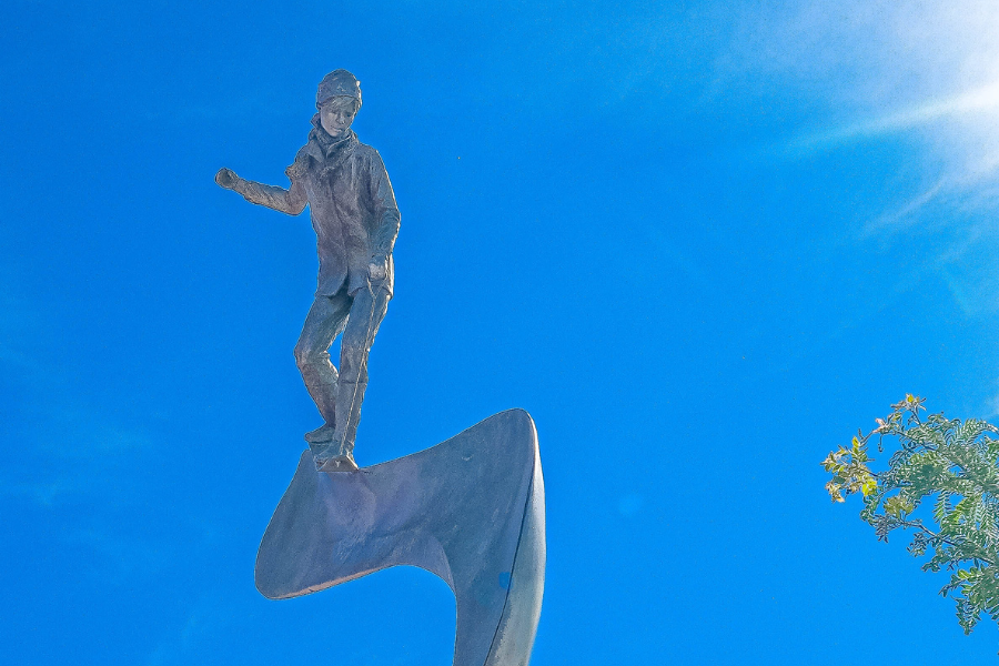 Statue of a person snurfing on a wave under a clear blue sky, with a small tree to the right.