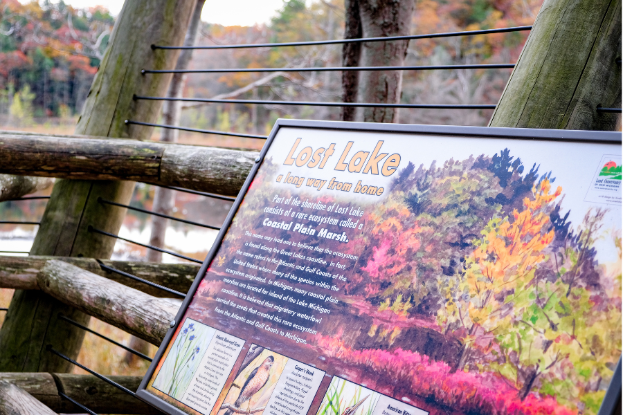 Interpretive “Lost Lake” sign displays autumn forest scenery and lake facts beside a rustic wooden overlook railing in a colorful woodland.