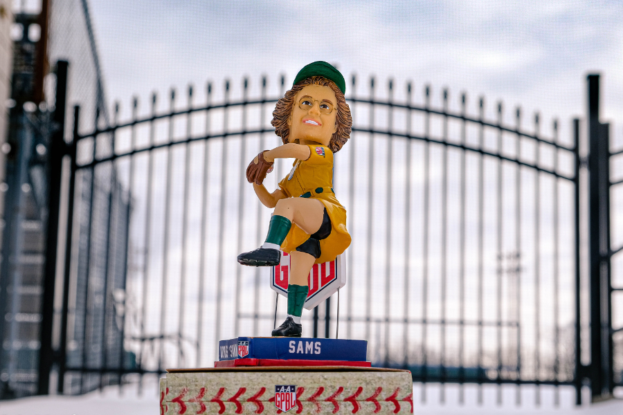 Bobblehead of the All-American Girls Professional Baseball League player Doris Sammy Sams from the perched on a pedestal with a metal fence in the background.