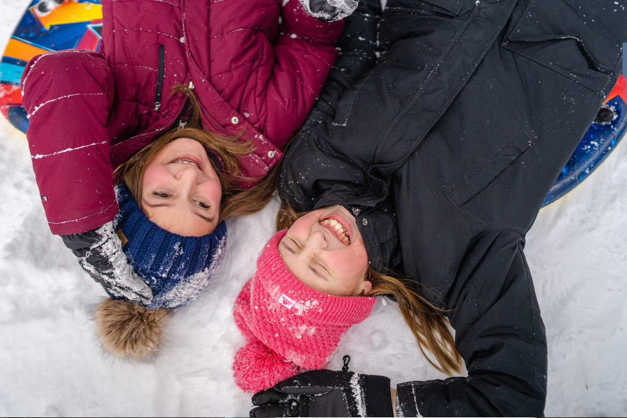 Two individuals lying in the snow, smiling and looking at the camera, with a colorful sled next to them.