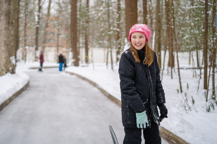 A person wearing a pink hat and black jacket is smiling while skating on a trail leading through a forest.