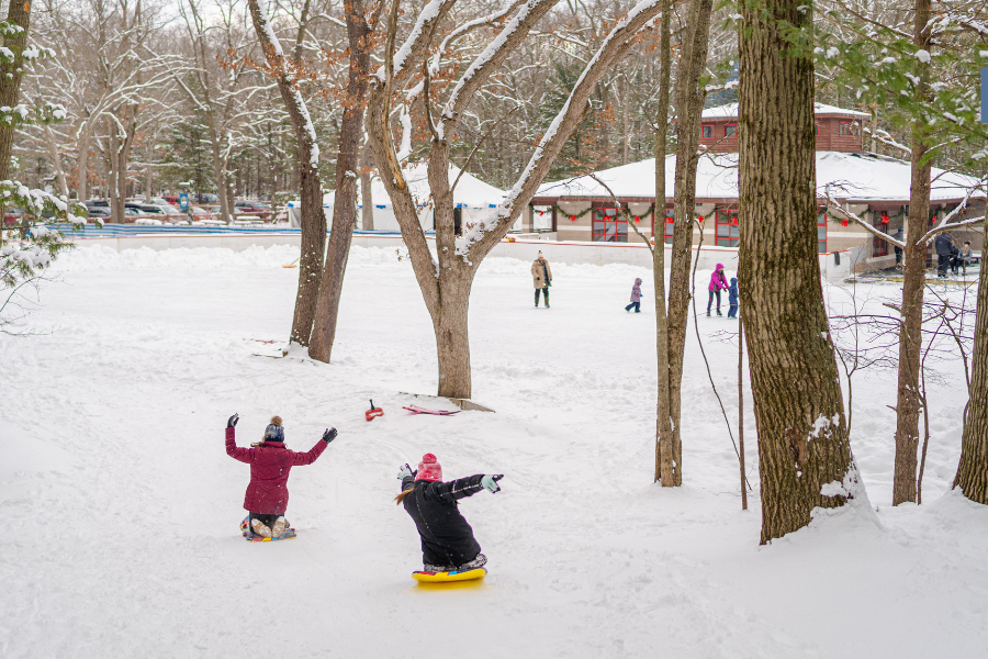 Two people sledding down a snowy hill at  Muskegon Luge Adventure Sports Park, with others visible in the background.