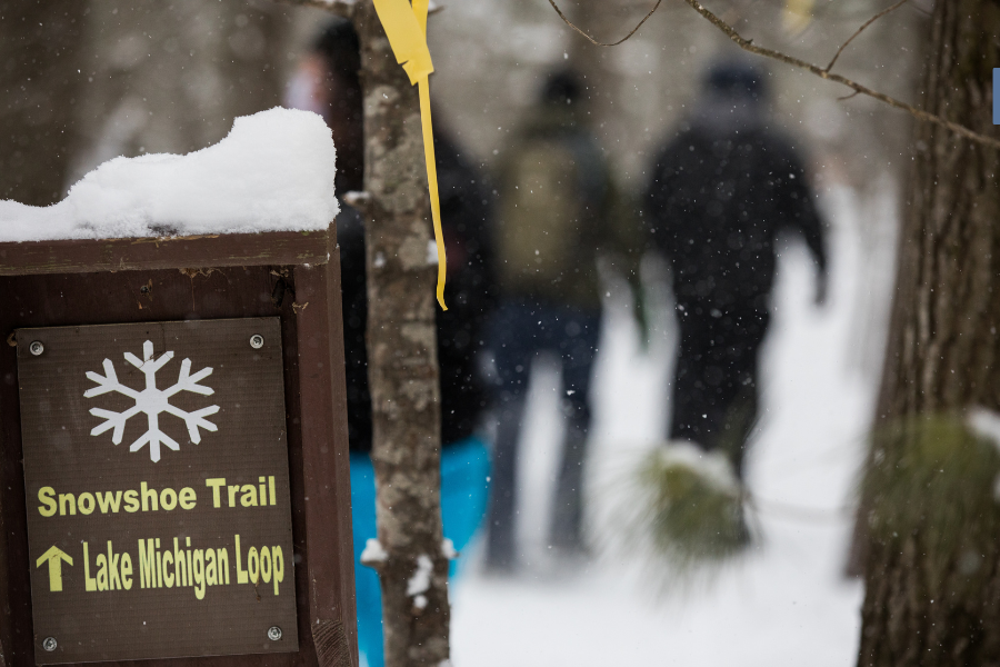 Sign indicating 'Snowshoe Trail Lake Michigan Loop' with a snow-covered top, beside blurred figures walking in the background through a snowy forest.