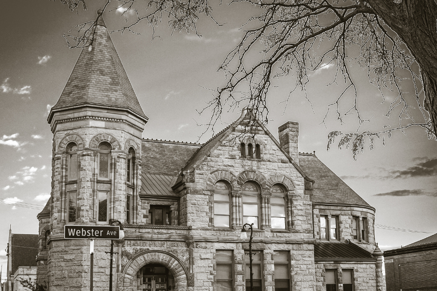 Sepia-toned image of the historic stone library building at the corner of Webster Ave, with a bare tree on the right and clear sky in the background.
