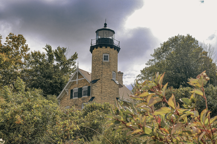 White River Light Station, a historic lighthouse and museum with a stone tower and attached keeper's house, surrounded by lush greenery under a cloudy sky.