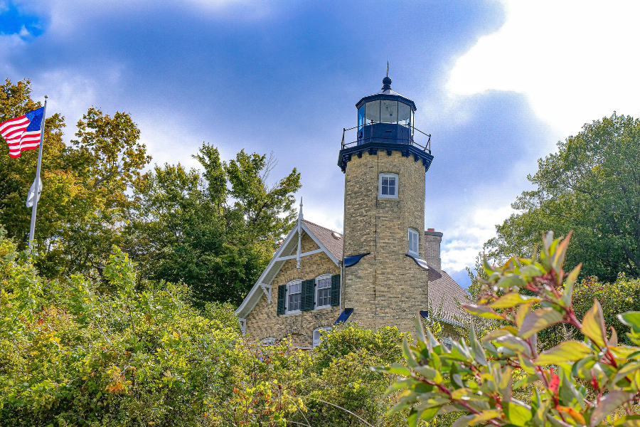 White River Light Station, a historical lighthouse with an attached light keepers house, under a partly cloudy sky with the American flag flying to the left, surrounded by lush greenery.
