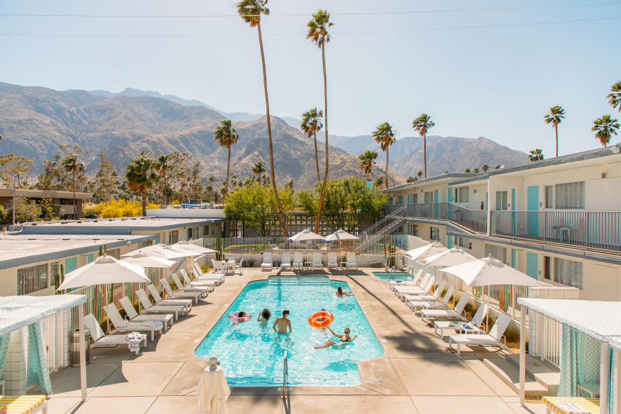An outdoor swimming pool at a hotel with people relaxing in and around the pool, surrounded by palm trees with mountainous scenery in the background. Sun loungers and umbrellas line the poolside, and a two-story hotel building flanks the pool area.