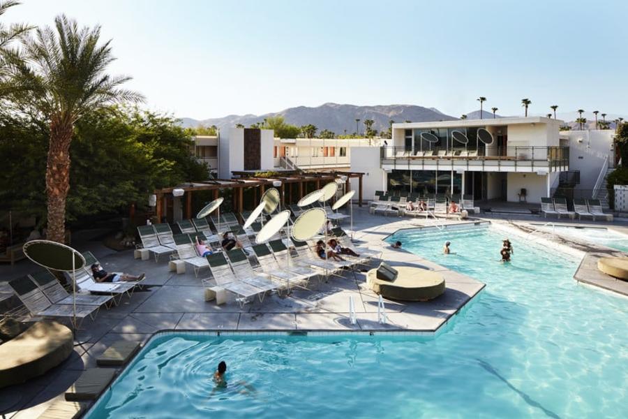 Outdoor swimming pool area at a modern hotel with people relaxing on sun loungers, palm trees, and mountainous background under a clear blue sky.