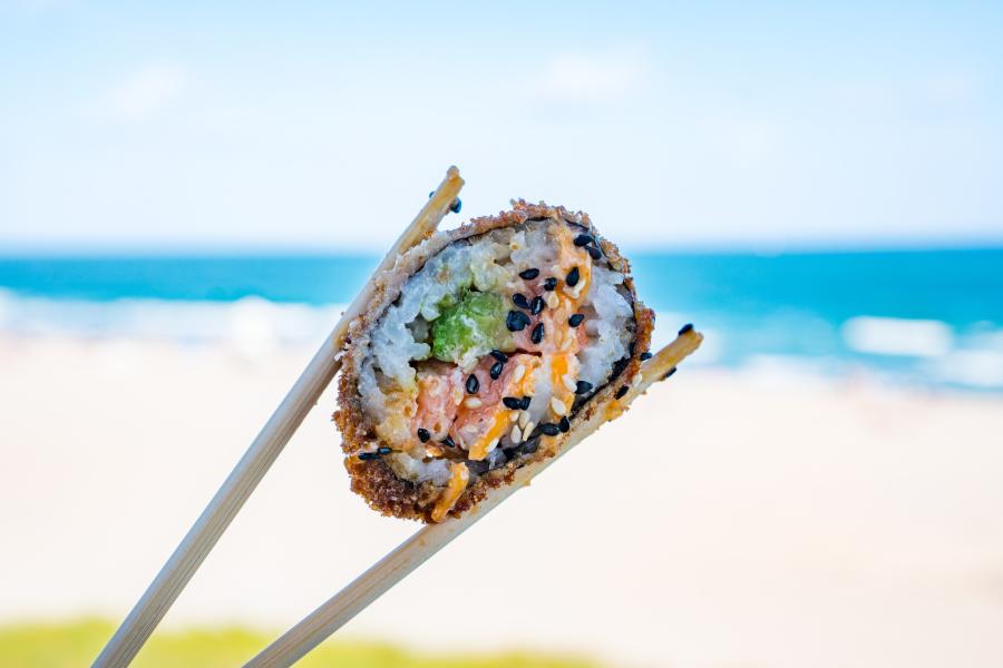 A piece of a sushi roll being held between two chopsticks with the ocean in the background