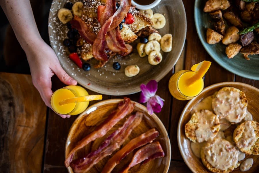 Aerial photo of four brunch dishes including a side of bacon, french toast with fresh fruit and bacon, hashbrowns, biscuits and gravy, and two mimosas with an orange slice garnish