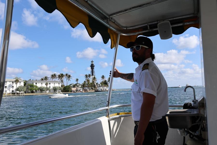 A uniformed boat captain standing aboard the Pompano Beach Water Taxi with the Hillsboro Lighthouse in the background