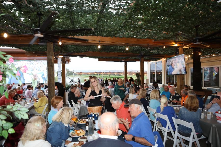 a covered patio with many tables filled with diners watching a large outdoor tv screen