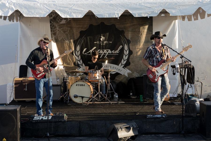 Three men in cowboy hats playing the guitar, bass guitar, and drums, on a small, covered stage in front of a banner that says "Old Town Untapped" in Pompano Beach.