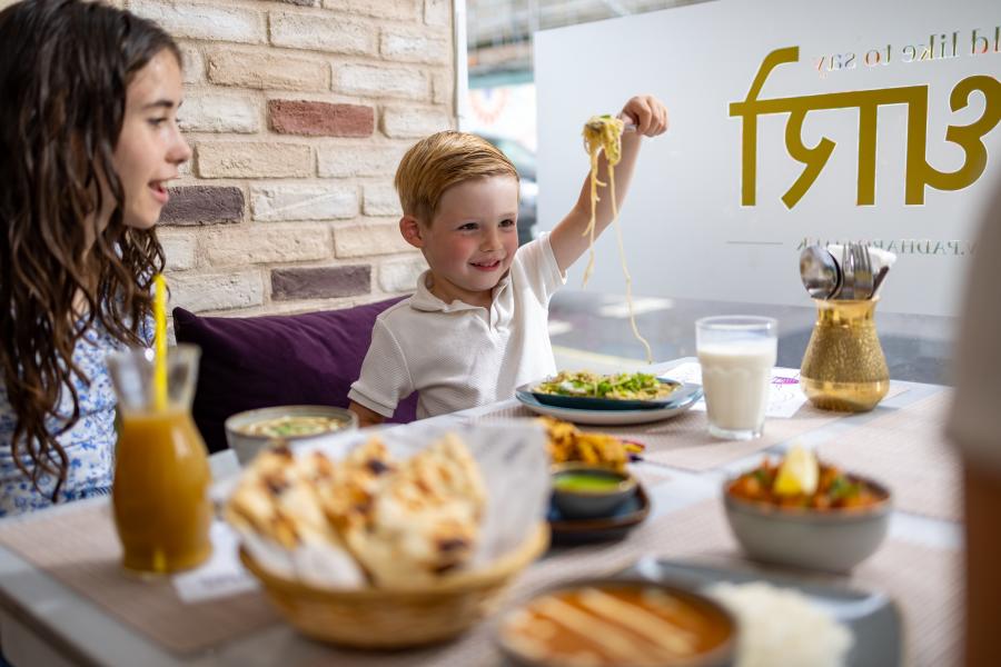 Child holding up noodles on a fork around table with family at Padharo