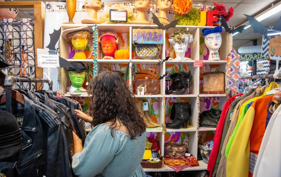 A girl with dark, curly hair looking at a rack filled with vintage clothes inside of a colorful thrift store.
