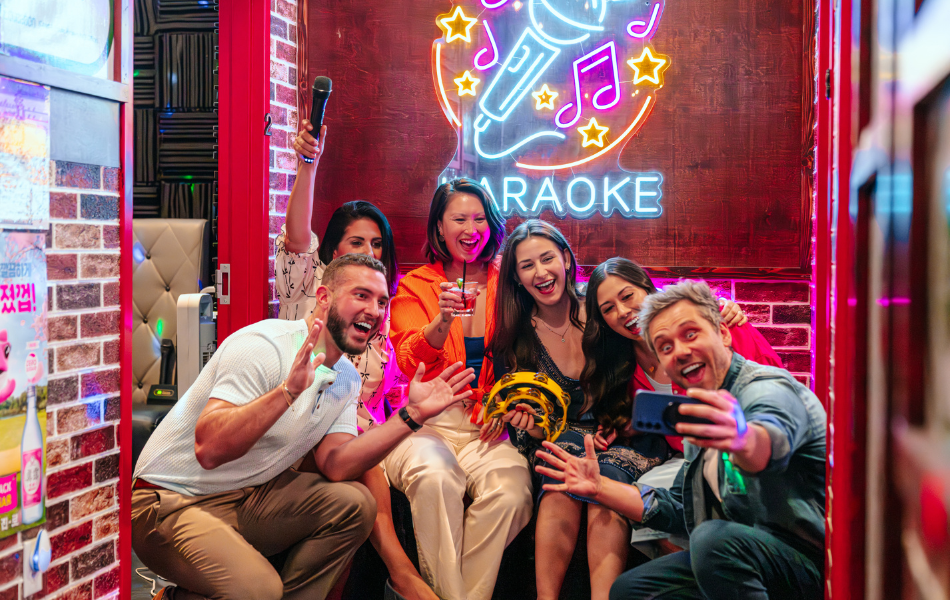 Group of friends smiling in front of a neon sign that reads "Karaoke"