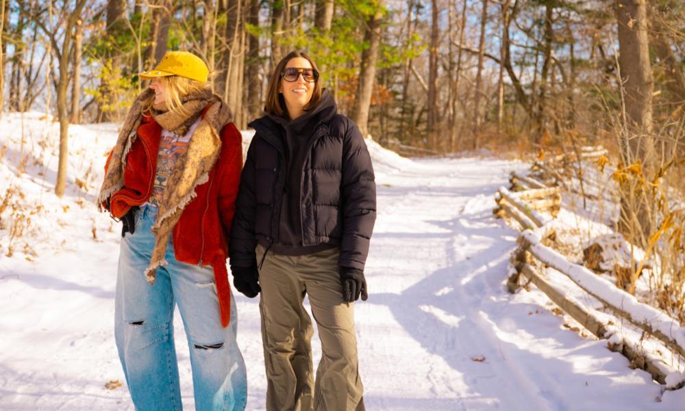 Two women stand on a snowy pathway outside surrounded by trees,  as they're holding hands and smiling while looking in opposite directions.