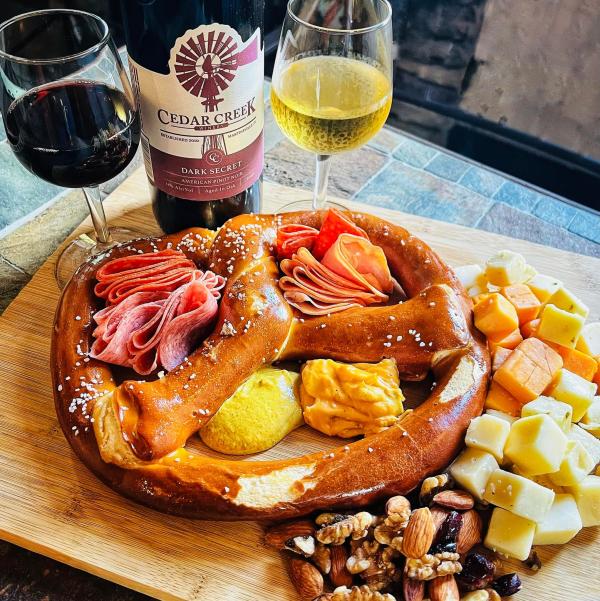 Image shows a pretzel charcuterie board from Cedar Creek Eatery in Martinsville, along with a glass of red wine and a glass of white wine. The large, soft pretzel is used to contain the various charcuterie items, including meats, cheeses, nuts, and mustard.