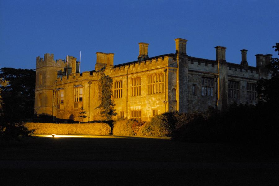 Sudeley Castle in the Cotswolds illuminated with warm golden lighting against a deep blue evening sky.