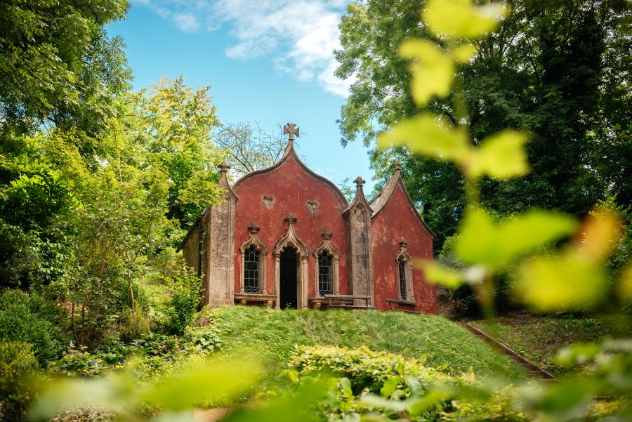 Historic red Gothic garden pavilion surrounded by lush greenery at Painswick Rococo Garden in the Cotswolds on a sunny day.