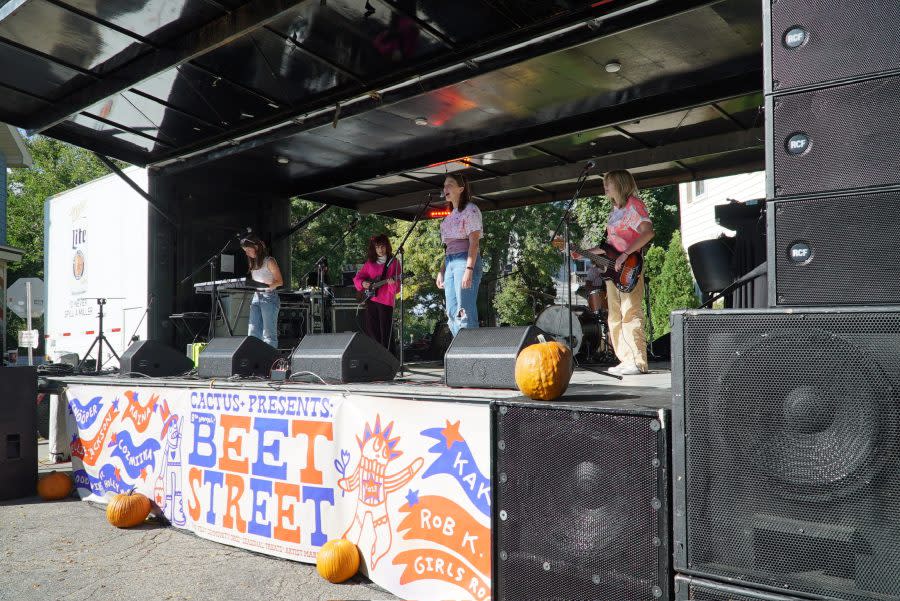 A band performs on an outdoor stage at Beet Street festival. Four musicians play keyboard, guitar, bass, and sing into microphones. The stage banner reads “Cactus Presents Beet Street” with colorful graphics, and pumpkins decorate the front of the stage.