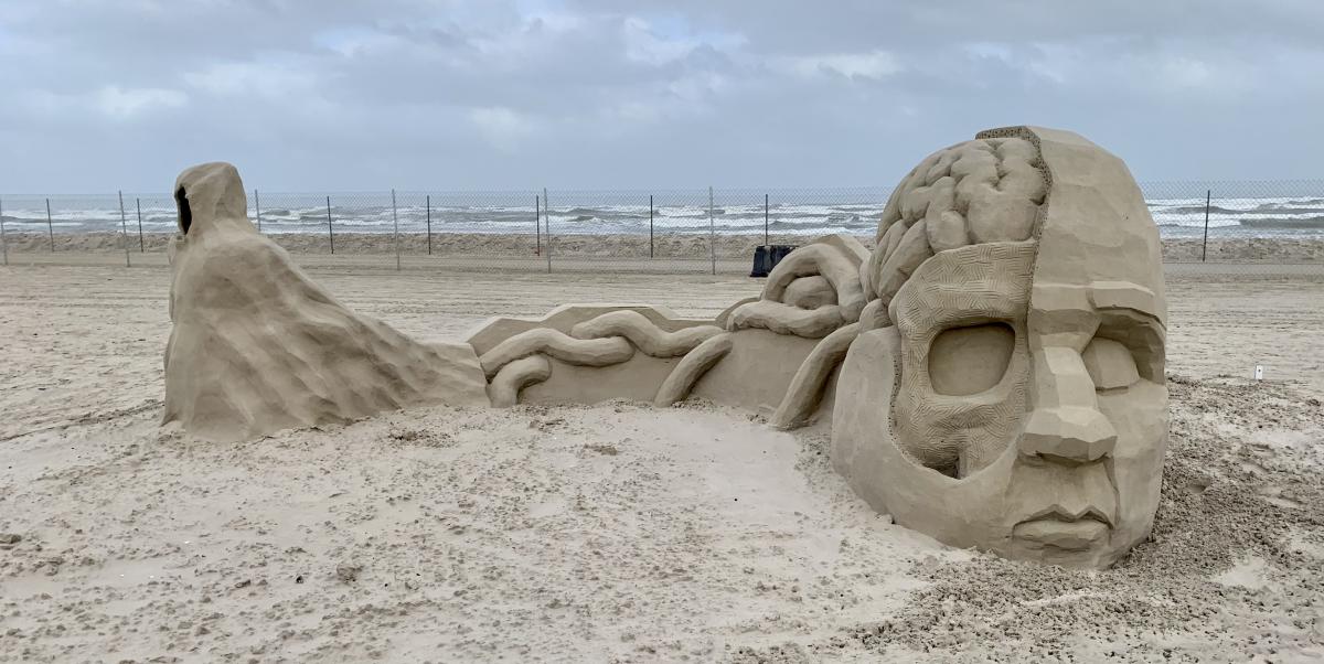 Sand sculpture showing a face on the right hand side, half exposed to the brain. On the left, the brain tendrils leak out to an ominous figure in the far left.