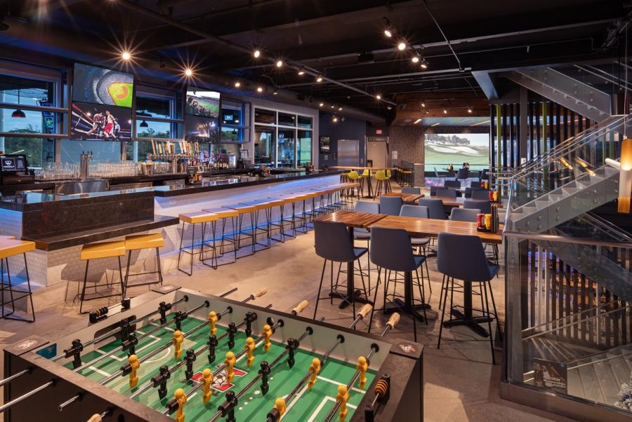 The third-level indoor area at Topgolf showing a long bar with seats along the front and TV screens behind it. There is also a foosball table, several high-top tables for dining, and a large-screen TV at the far end.