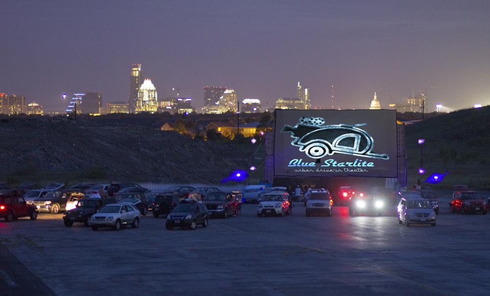 Cars backed up to screen at Blue Starlite Mini Urban Drive-In, with illuminated skyline in the background.