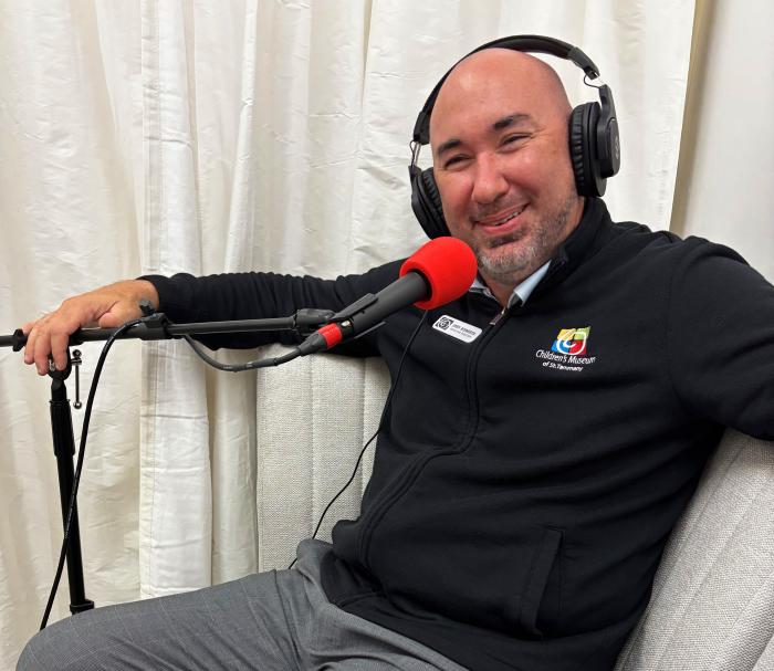 Joey Roberts, Executive Director of the Children's Museum of St. Tammany, smiles during an All The Waves podcast recording. Wearing headphones and a Children's Museum logo jacket, he sits in a chair speaking into a microphone while discussing the museum's programs and impact on local families.