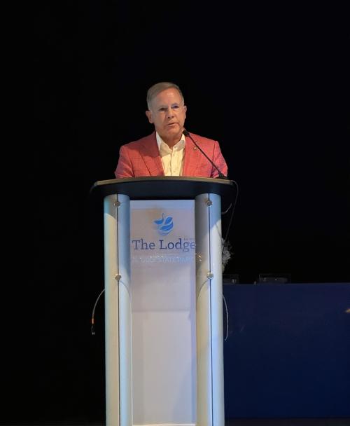 A businessman in a red coat and white button down speaks from behind a podium