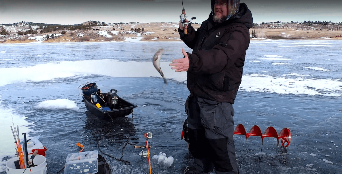 A man ice fishing at Curt Gowdy State Park, holding a fishing rod with a fish leaping out of the water, surrounded by ice fishing equipment and a snowy landscape in the background.