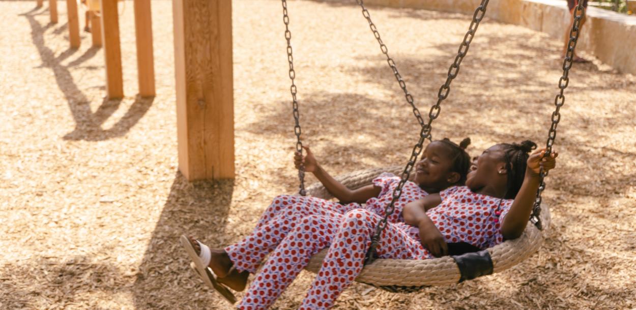 Two kids laughing on swing together in matching outfits at a local park.