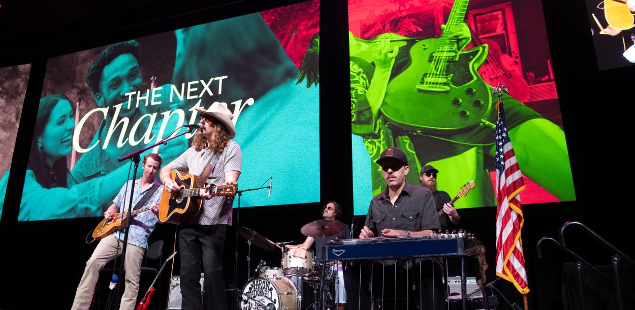 Band playing instruments on a stage with a large screen that says "
