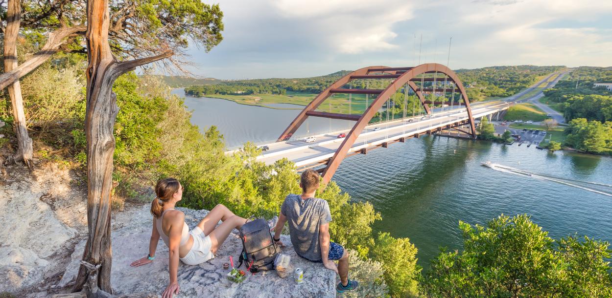 Hikers sitting on a hilltop rock overlooking the Pennybacker Bridge at Lake Austin.