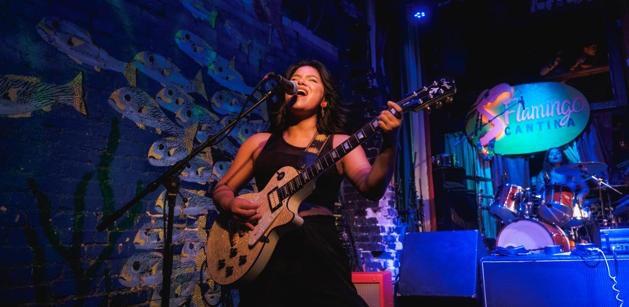 Woman standing on a stage painted with an irredescent fish backdrop, playing the electric guitar and singing into a microphone.