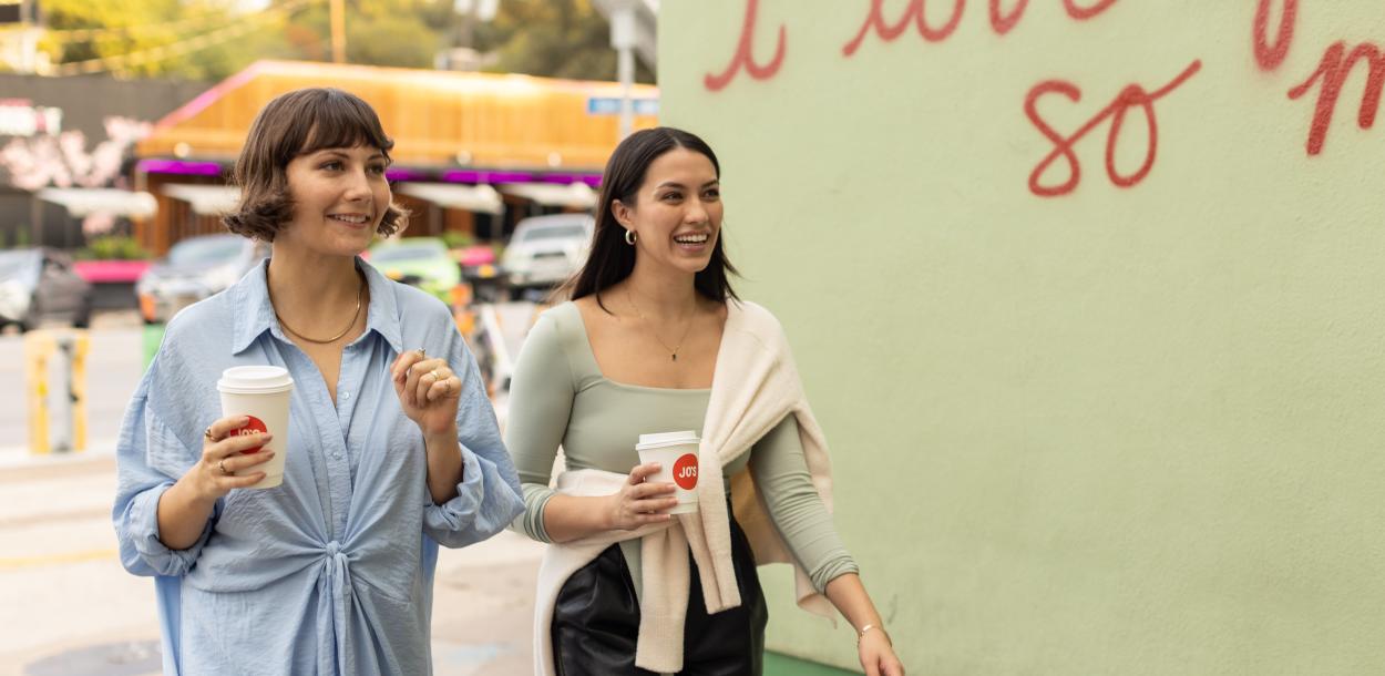 Two women walking past "I love you so much." mural with to-go coffee cups in hand.