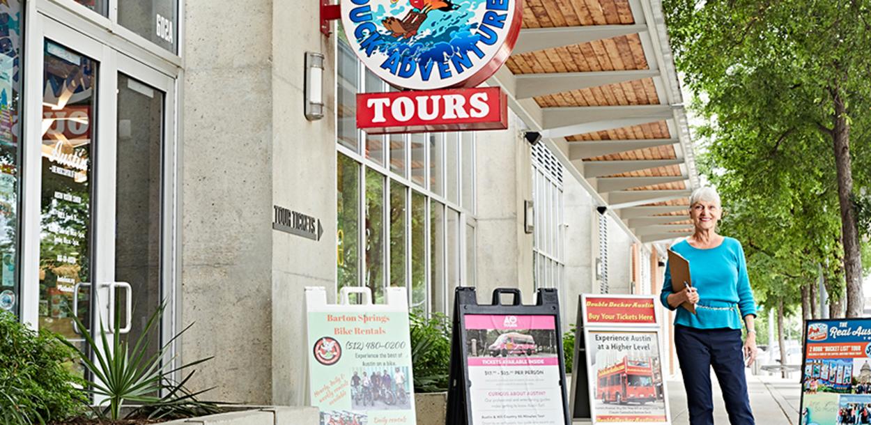 White haired woman stands outside the Austin Visitor Center with a clipboard. Behind her, there are multiple sandwich board signs promoting local tours