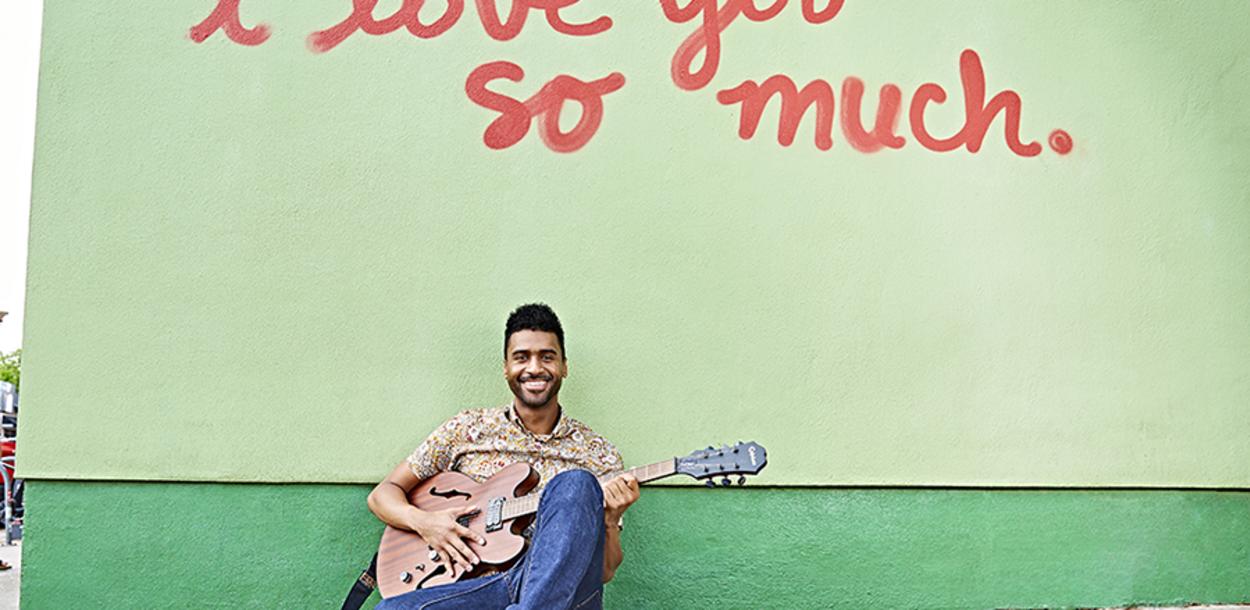 Musician Tje Austin sits below a mural reading "I love you so much." on South Congress Ave. He is holding a guitar and has a cup of coffee sitting next to him