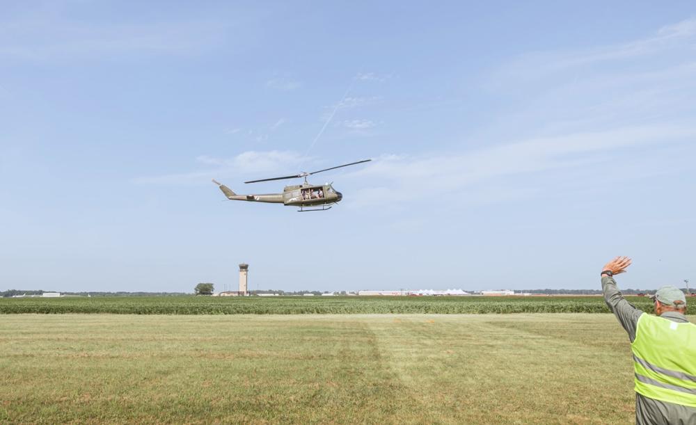 Helicopter takes off from grassy field. Person in right corner of photo standing in yellow safety vest, waving goodbye to the plane passengers.
