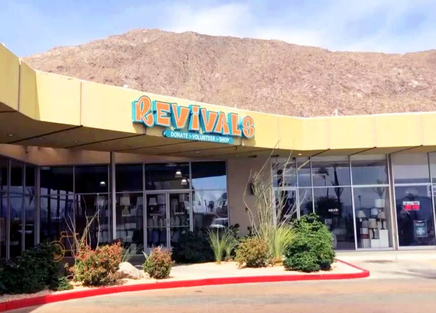A storefront with the sign 'REVIVALS' above the entrance, surrounded by a sunny day, sparse vegetation, and a large hill in the background.