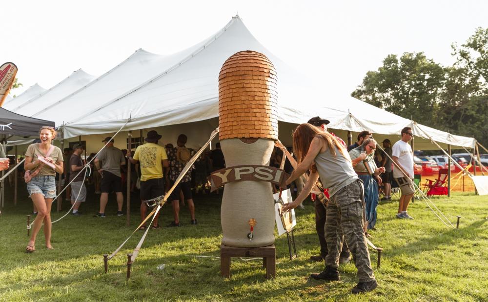 Man with long hair visits the beer tap shaped like the Ypsilanti Water Tower with the word 'Ypsi' written across the front. The man pours a beer into his cup from the tap.