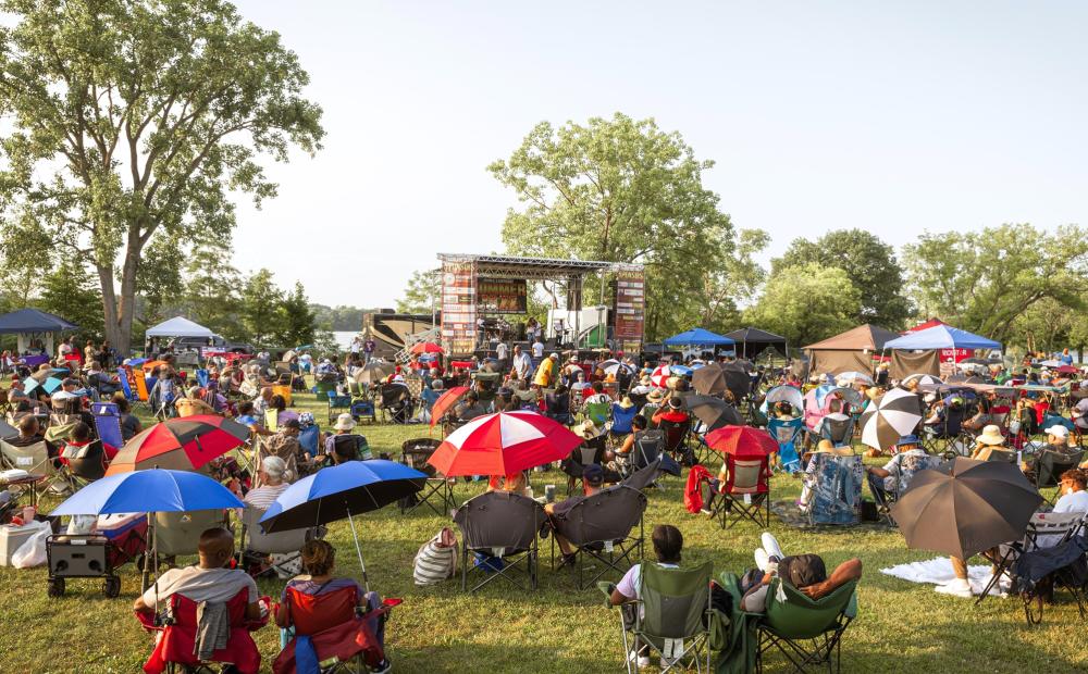 Large crowd with lawn chairs and propped up umbrellas outdoors in a park with green grass and trees,. Crows faces the stage in the distance, as a musician performs music on stage with a flute.