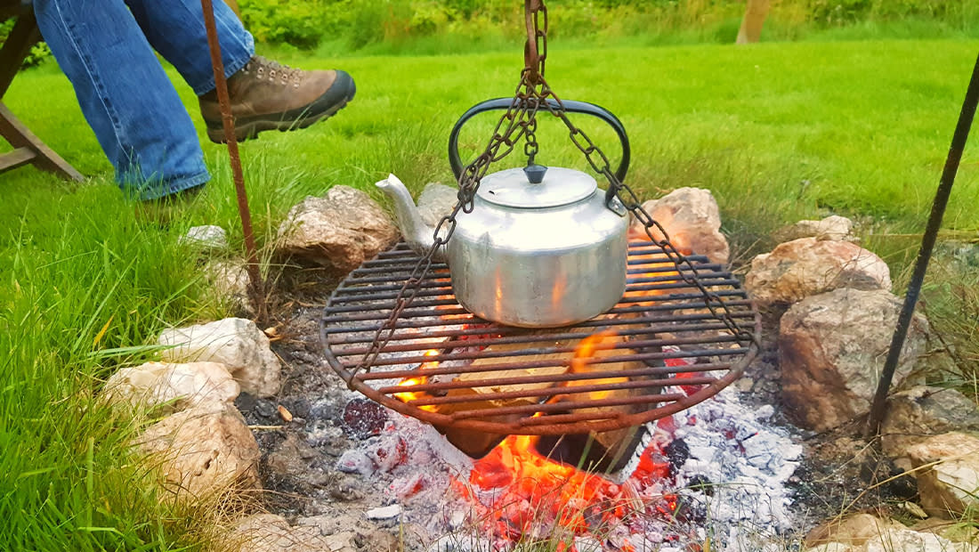 A kettle hangs over a crackling campfire on a metal grill, surrounded by rocks and lush grass. A person in jeans and boots sits nearby, evoking a calm outdoor setting