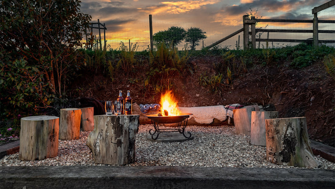 A cosy outdoor setting with a central fire pit surrounded by tree stump stools. Two beer bottles and glasses sit nearby. The sunset sky adds warmth.