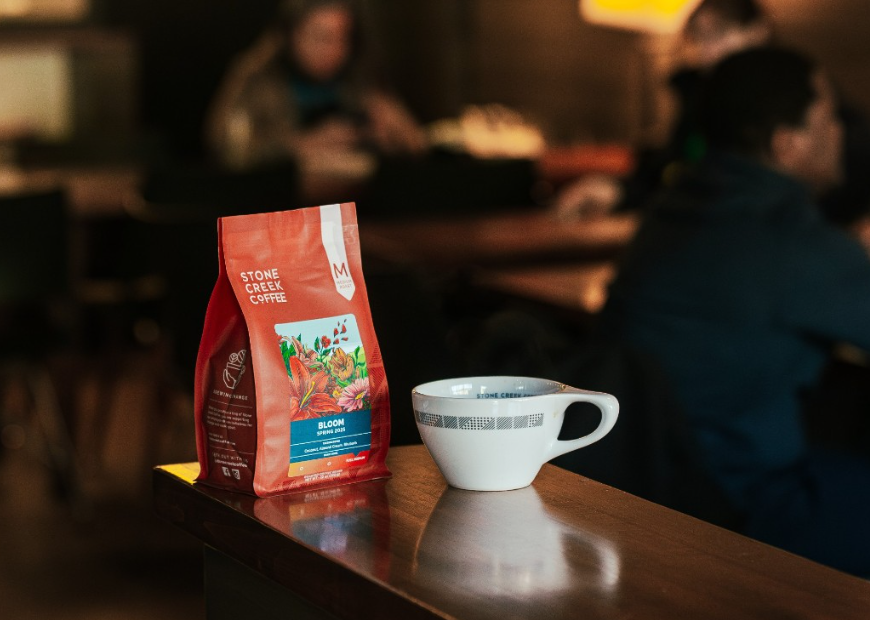 Bag of Stone Creek Coffee and a white ceramic coffee cup on a wooden counter, with a blurred café setting in the background.
